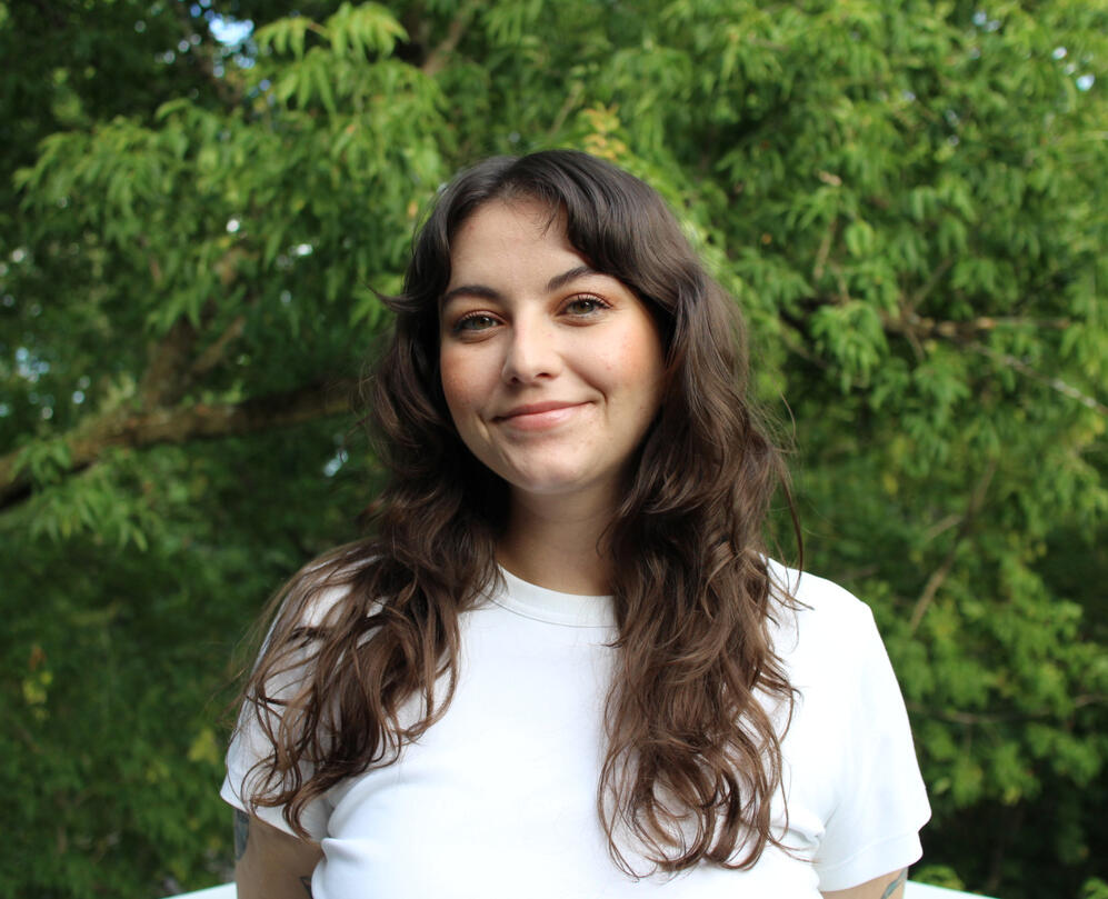 Kendal smiling softly. She is white, has long wavy brown hair, green eyes, and is wearing a white t-shirt. There are leafy green trees in the background.
