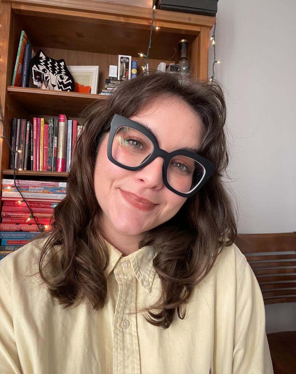 Kendal smiling softly for a selfie. She has medium length brown wavy hair, and is wearing bold oversized black glasses. She is wearing a butter yellow button-up shirt, buttoned all the way up.